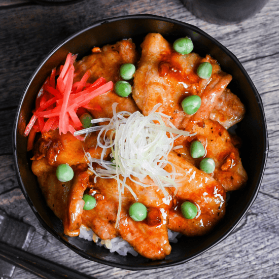 Japanese pork bowl (butadon) made with caramelized pork belly slices, green peas, pink pickled ginger and strings of Japanese leek in a black bowl on a wood-effect background