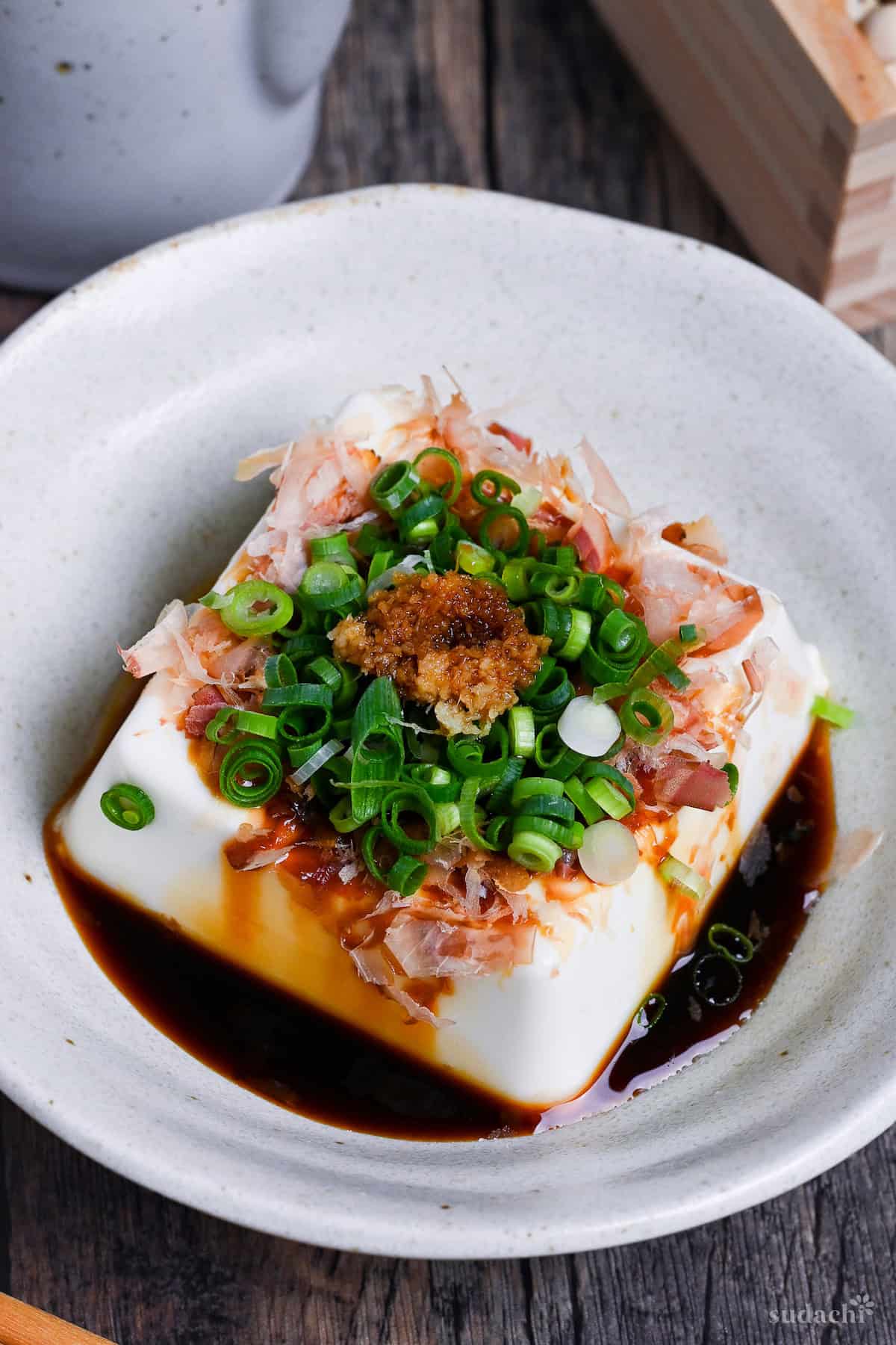 Hiyayakko, a block of silken tofu topped with soy sauce, bonito flakes, green onions and grated ginger in a white ceramic dish on a wooden background close up