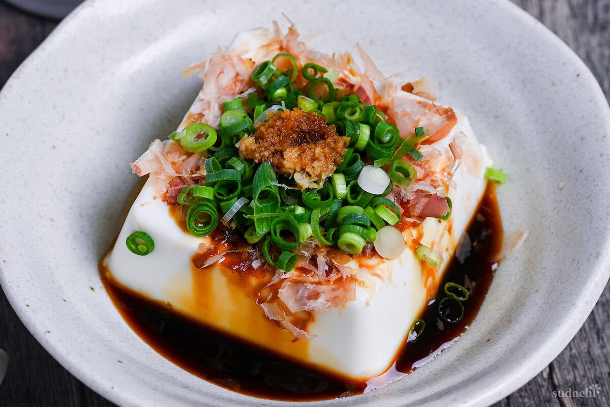 Hiyayakko, a block of silken tofu topped with soy sauce, bonito flakes, green onions and grated ginger in a white ceramic dish on a wooden background