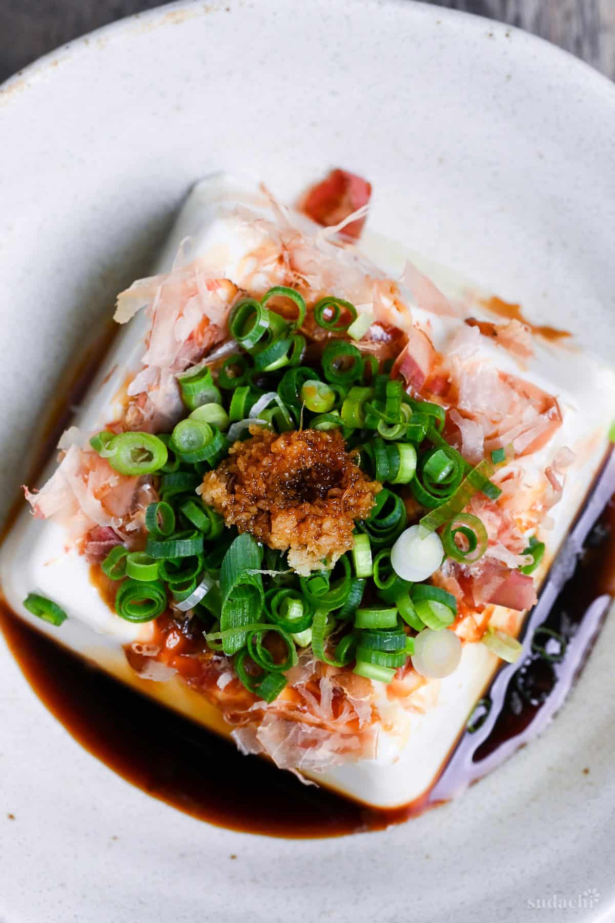 Hiyayakko, a block of silken tofu topped with soy sauce, bonito flakes, green onions and grated ginger in a white ceramic dish on a wooden background top down close up
