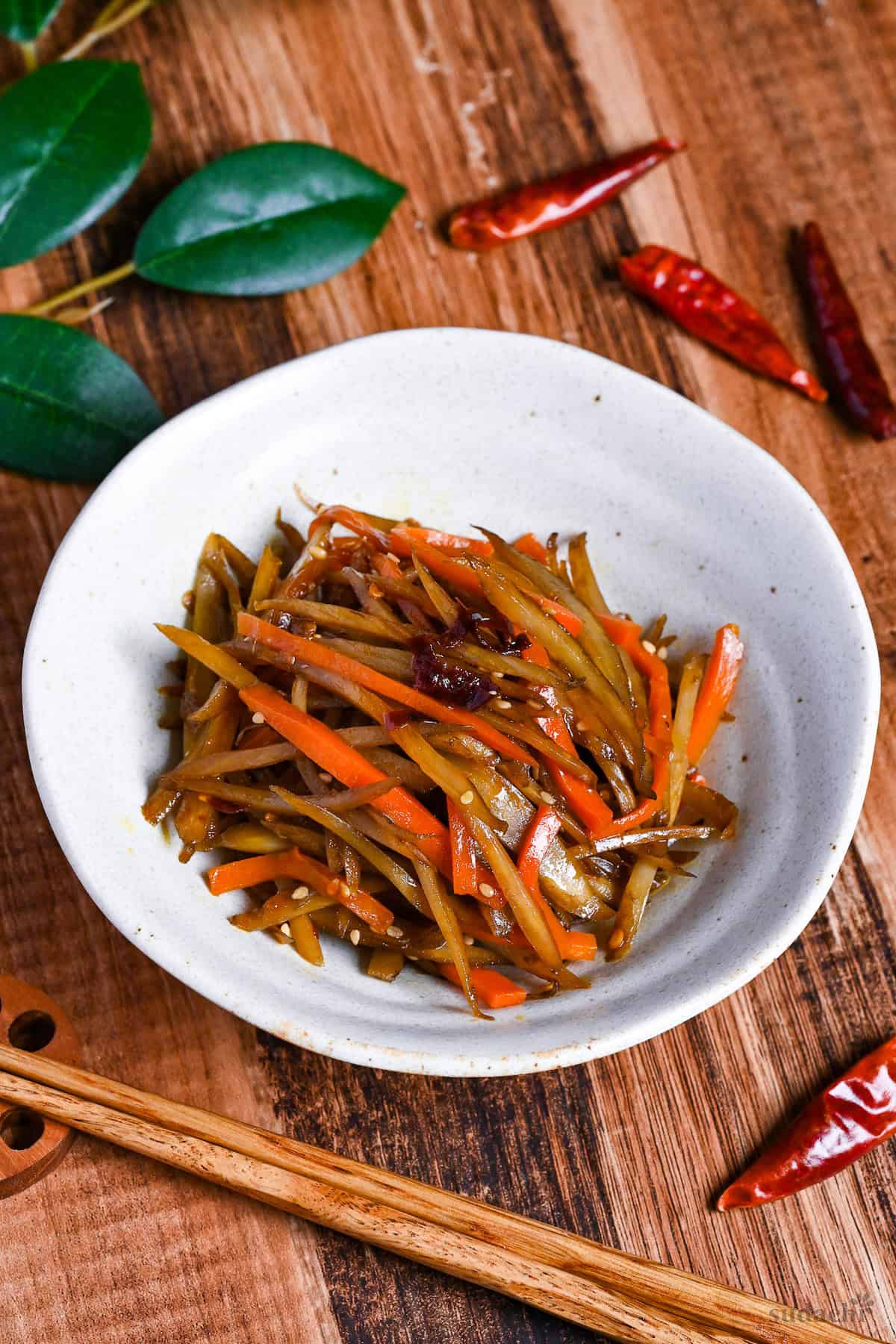 Kinpira gobo made with carrots and burdock root topped with red chili in a white bowl on a wooden background surrounded by dry red chilis