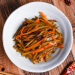 Kinpira gobo made with carrots and burdock root topped with red chili in a white bowl on a wooden background surrounded by dry red chilis