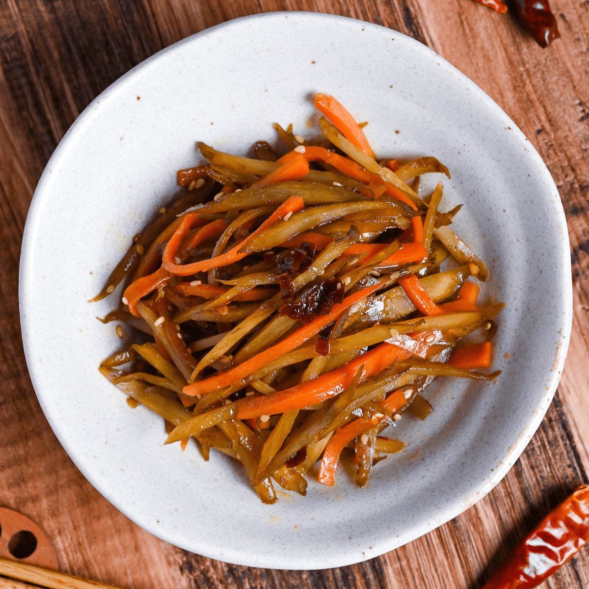 Kinpira gobo made with carrots and burdock root topped with red chili in a white bowl on a wooden background surrounded by dry red chilis