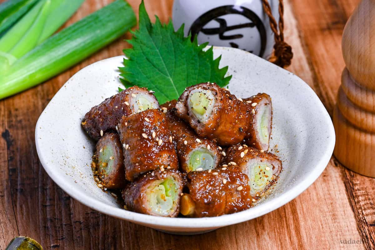 Glazed beef negimaki (beef leek rolls) cut into bitesize pieces in a white bowl decorated with a shiso leaf and sprinkled with shichimi togarashi and sesame seeds on a wooden background