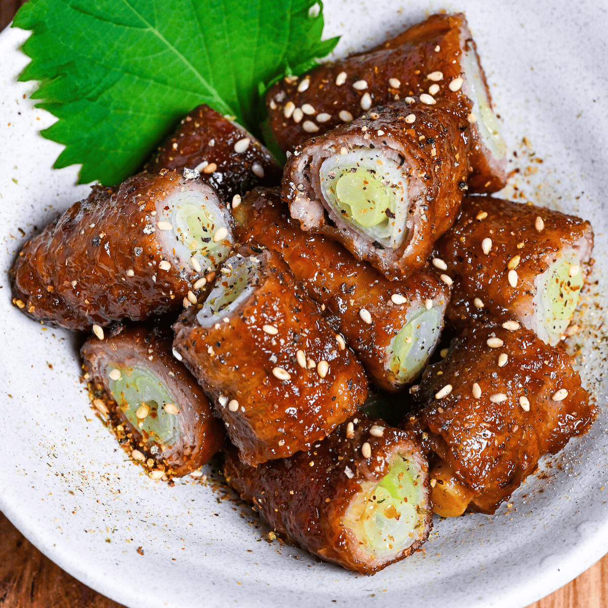 Glazed beef negimaki (beef leek rolls) cut into bitesize pieces in a white bowl decorated with a shiso leaf and sprinkled with shichimi togarashi and sesame seeds on a wooden background