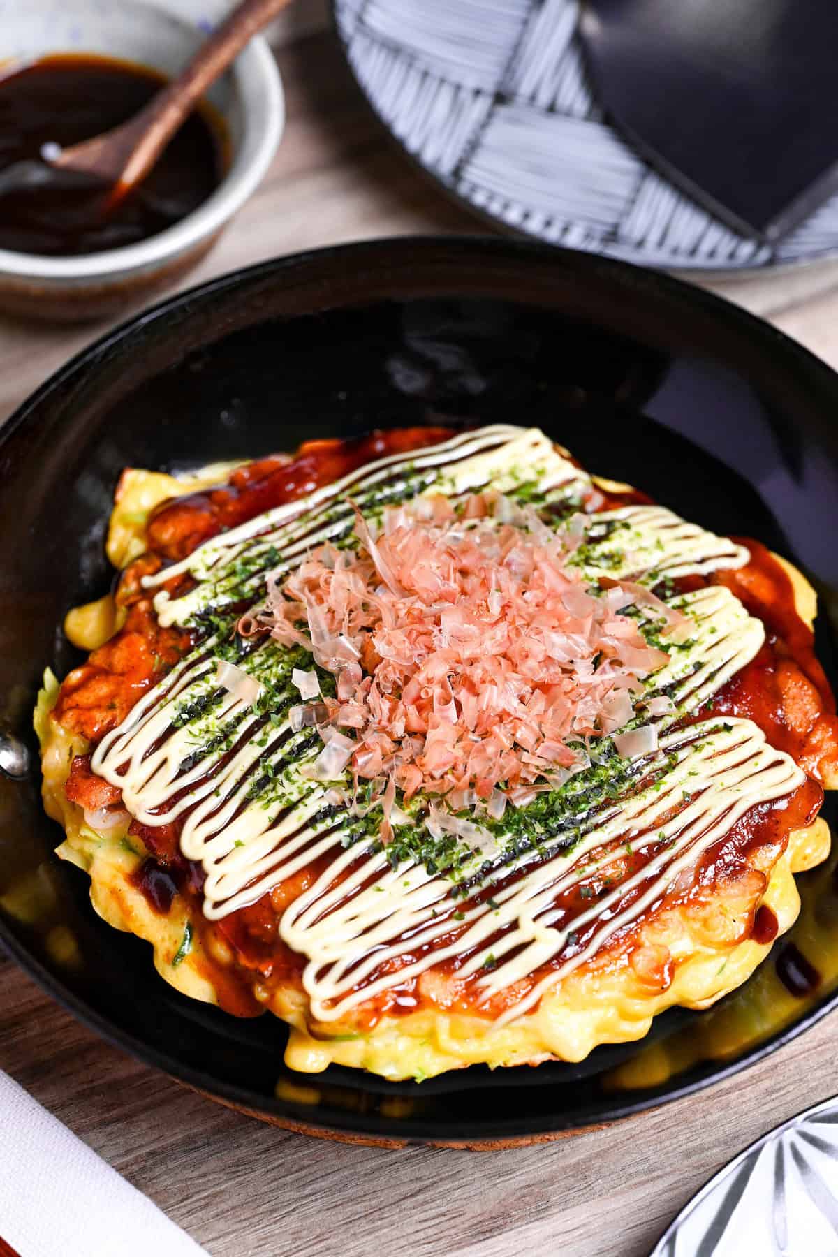Osaka style okonomiyaki topped with homemade okonomi sauce, mayonnaise, aonori and bonito flakes in a small frying pan with sauce and a plate in the background