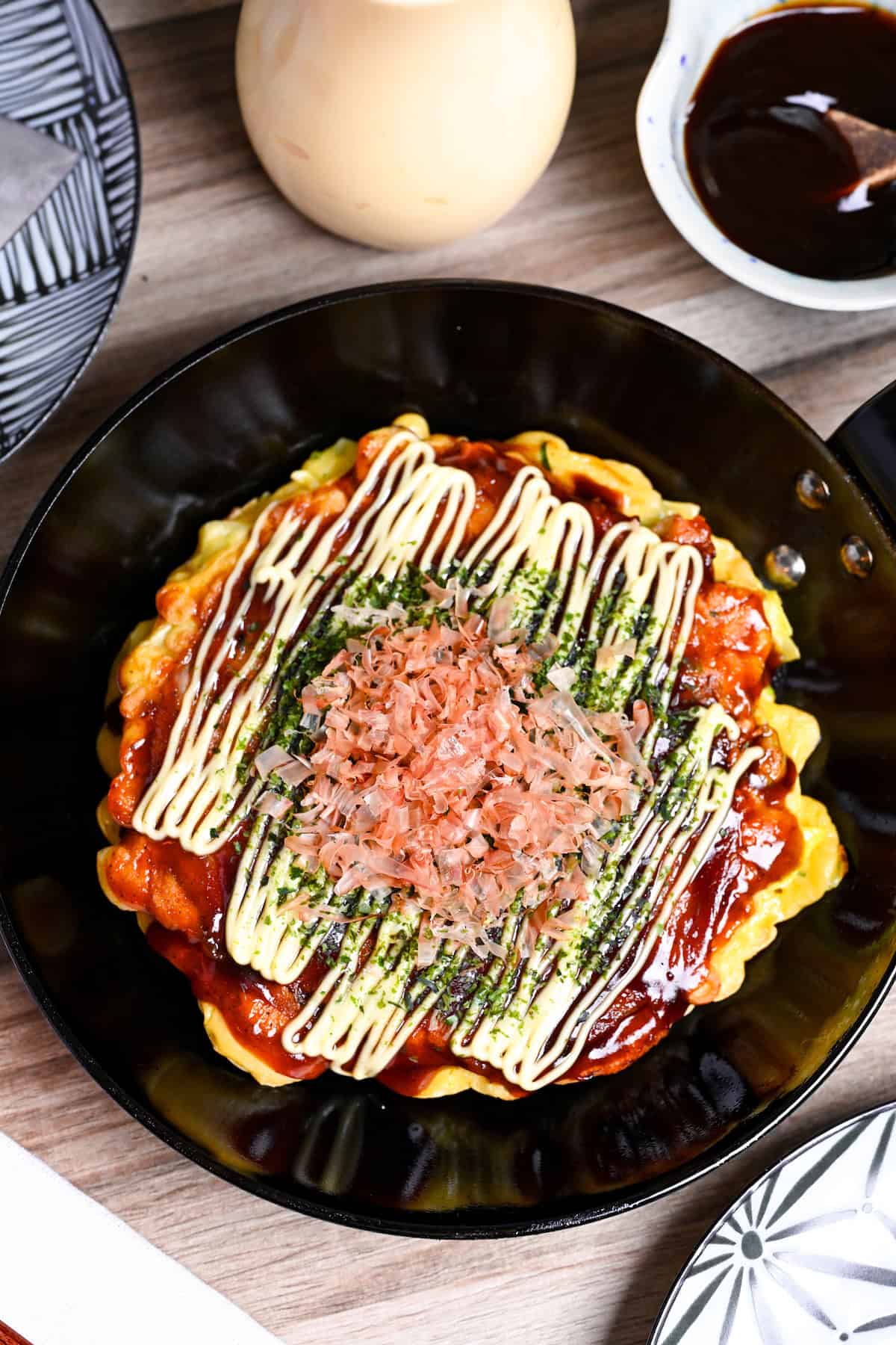 Osaka style okonomiyaki topped with homemade okonomi sauce, mayonnaise, aonori and bonito flakes in a small frying pan with sauce and a plate in the background