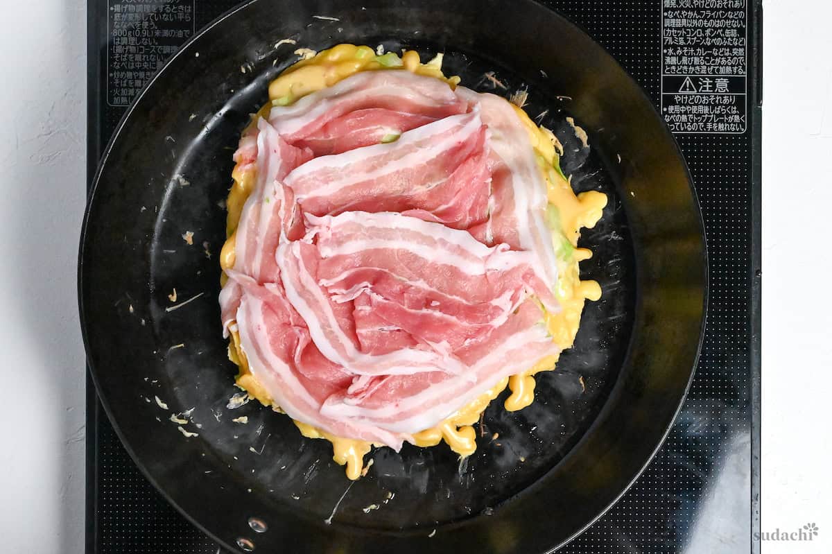 okonomiyaki topped with thinly sliced pork belly in a cast iron frying pan on the stove