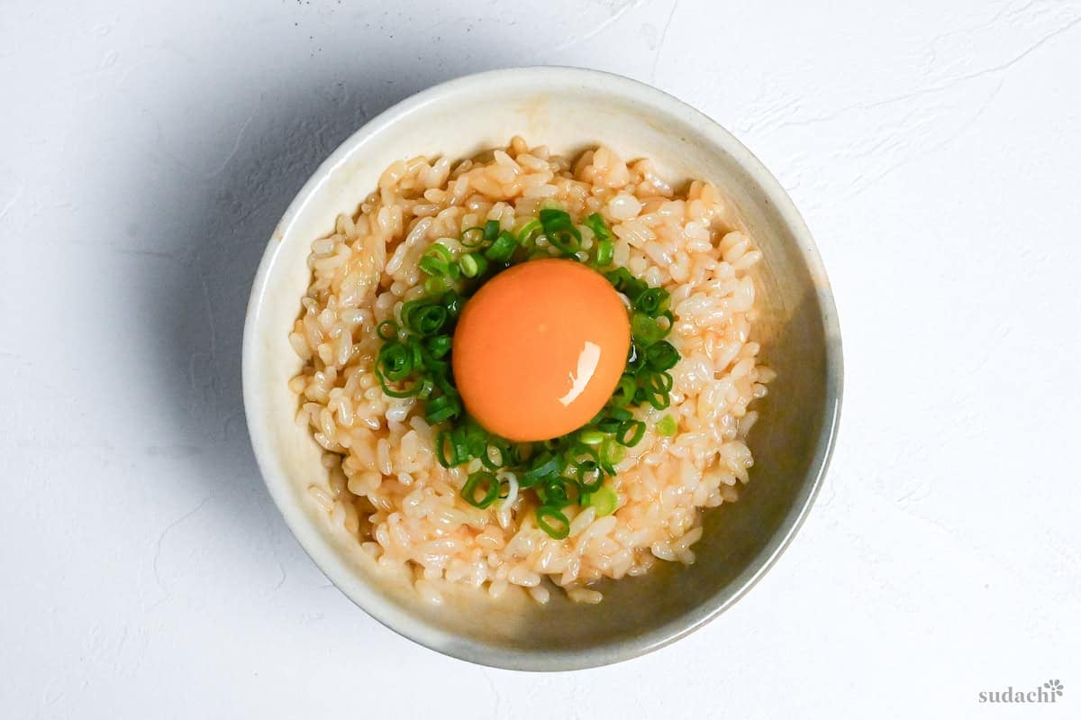 Tamago kake gohan topped with chopped green onions and a raw egg yolk in a cream ceramic bowl on a white background