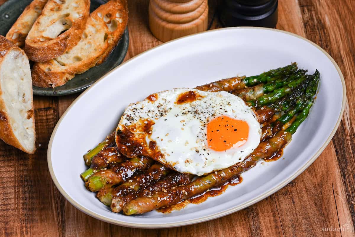 Side view of nikumaki (pork belly wrapped) asparagus on a white plate topped with sunny side up egg with sliced toasted baguette and salt and pepper grinders in the background