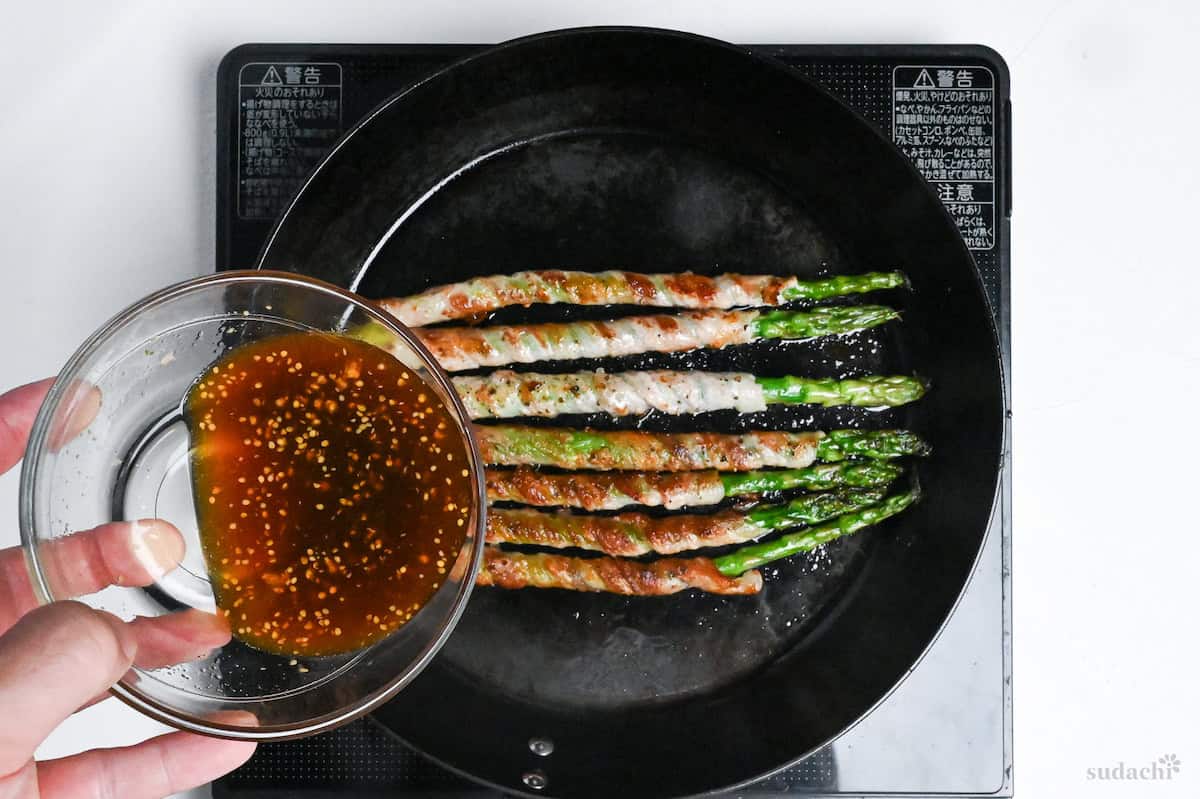 Pouring sauce over nikumaki (pork belly wrapped) asparagus in a frying pan on the stove top