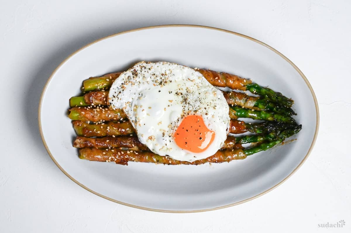Nikumaki (pork belly wrapped) asparagus topped with sunny side up egg and freshly ground black pepper on a white oval plate