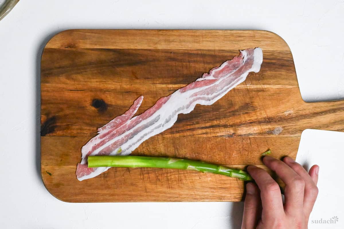 Asparagus stalk placed at 90 degrees over a strip of thinly sliced pork belly placed at 45 degrees on a wooden chopping board