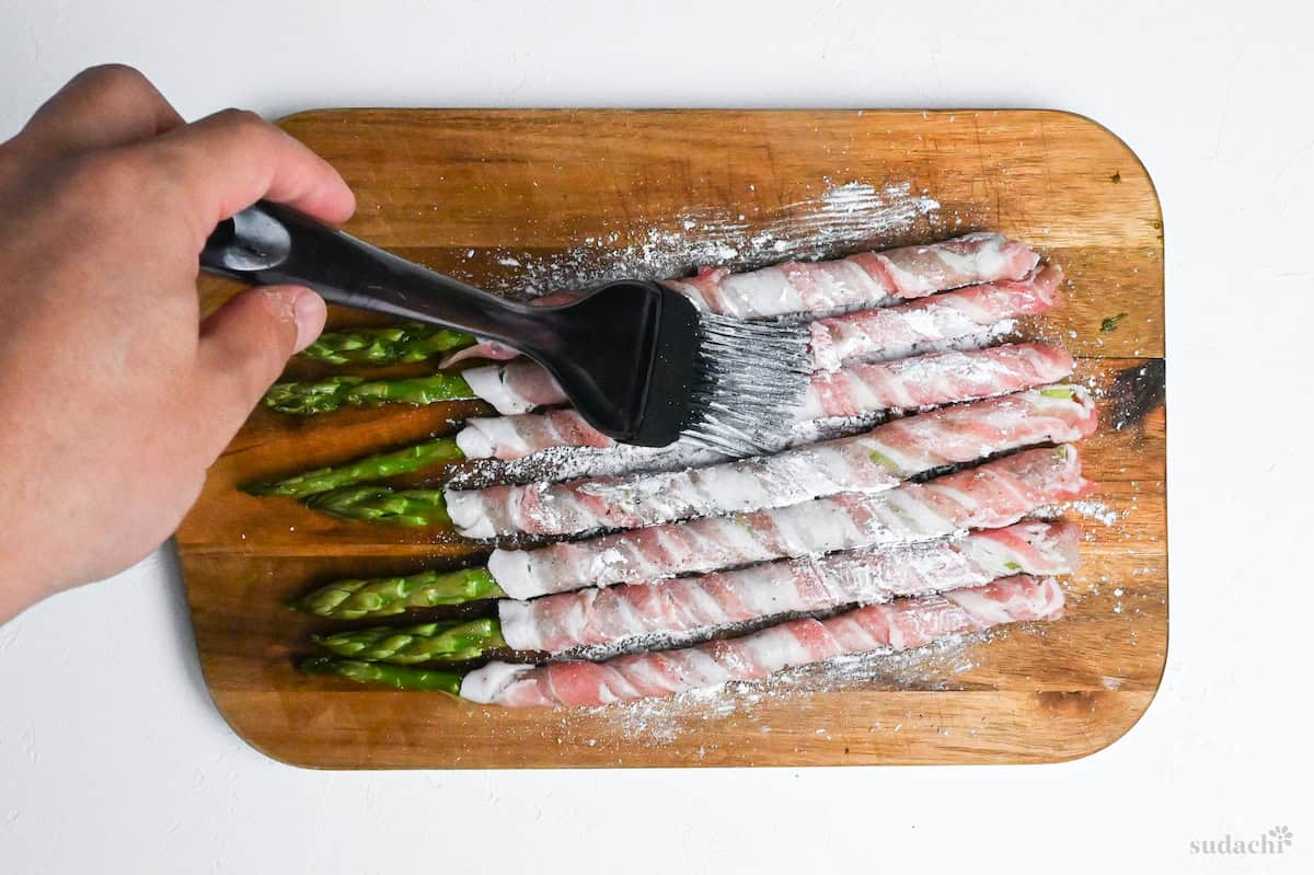 Brushing pork wrapped asparagus with cornstarch on a wooden chopping board on a white background