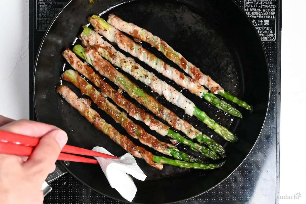 Holding kitchen paper with red cooking chopsticks to wipe out excess fat in pan when frying nikumaki (pork belly wrapped) asparagus