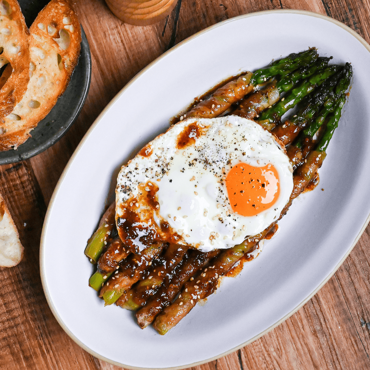 Nikumaki (pork belly wrapped) asparagus topped with sunny side up egg and sprinkled with freshly ground black pepper on a white plate on a wood-effect background