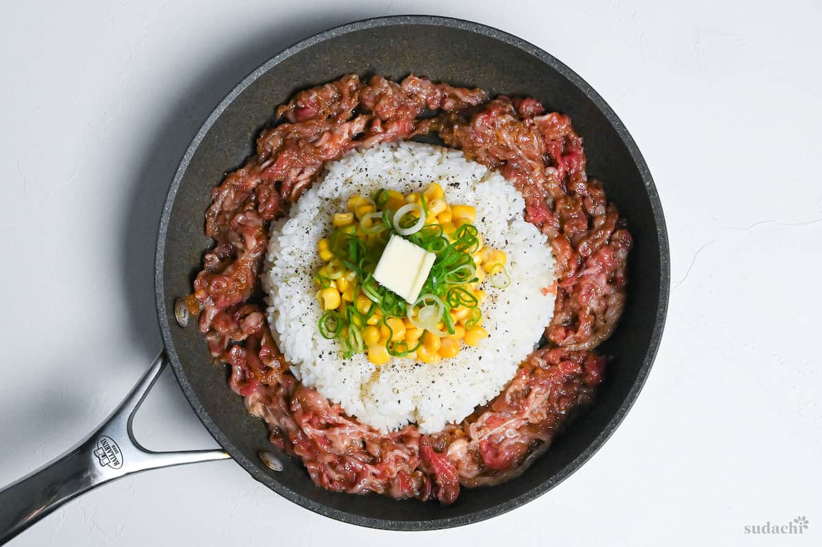 cooked rice in a frying pan topped with sweet corn, green onions and a pat of butter surrounded by thinly sliced marinated beef