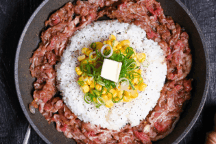 Beef pepper rice in a frying pan on a black wood effect background surrounded by a block of butter, a bowl of sauce and a wooden pepper grinder