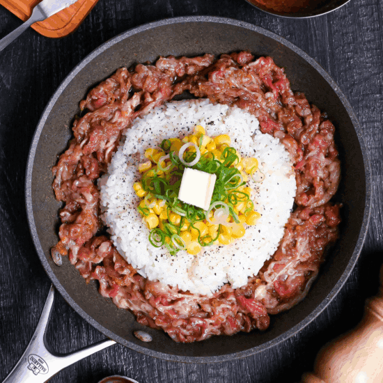 Beef pepper rice in a frying pan on a black wood effect background surrouneded by a block of butter, a bowl of sauce and a wooden pepper grinder