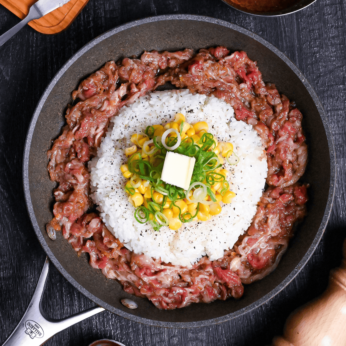 Beef pepper rice in a frying pan on a black wood effect background surrounded by a block of butter, a bowl of sauce and a wooden pepper grinder