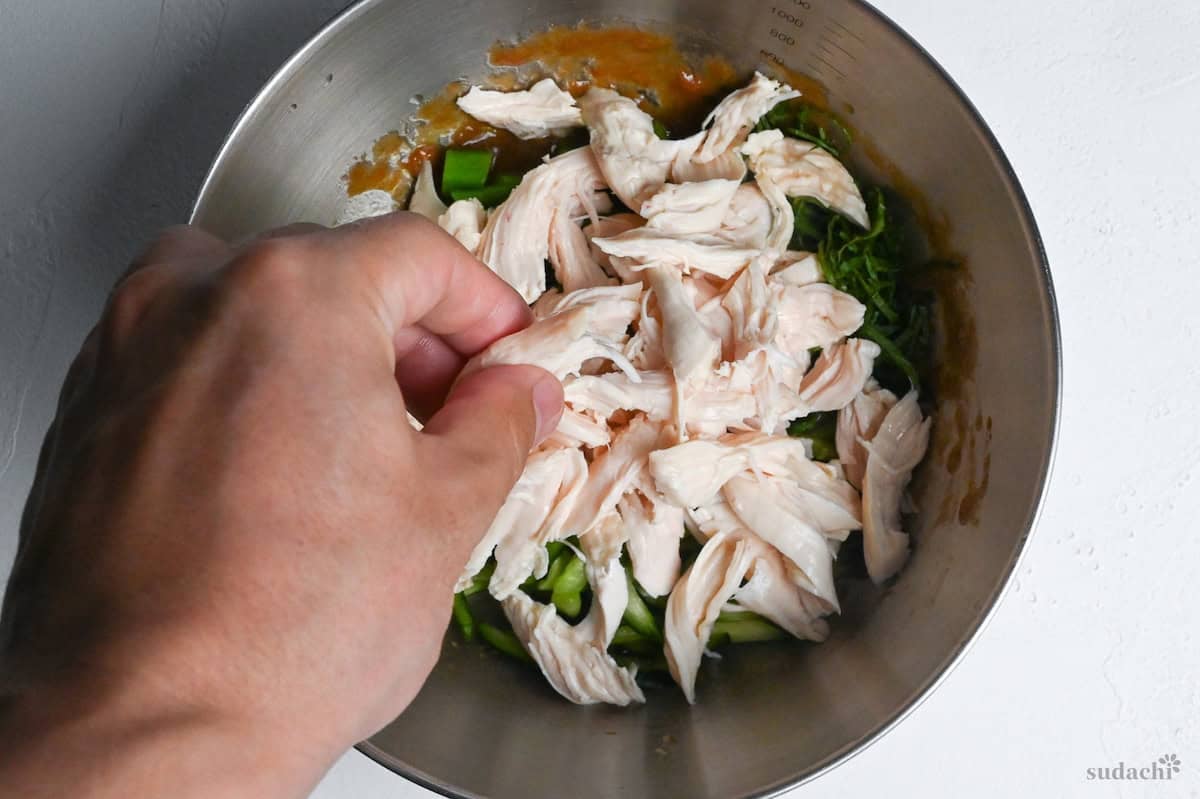 shredding poached chicken tenders into the bowl of dressing and vegetables to make salad