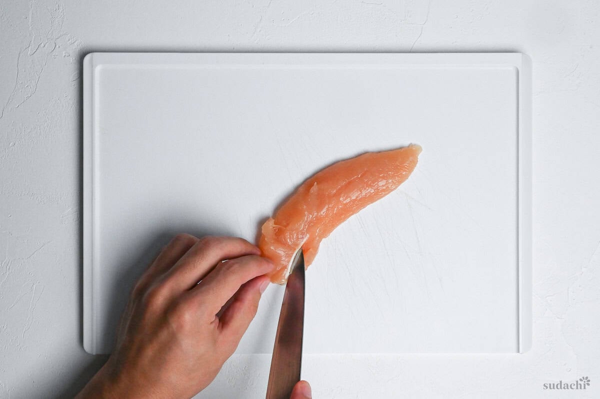 cutting the tendon in one chicken tenderloin on a white cutting board
