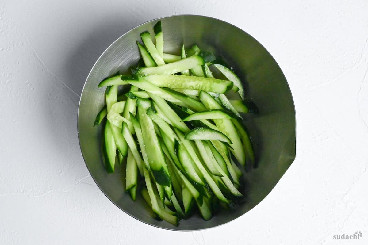 julienned cucumber salted in a steel mixing bowl on a white background