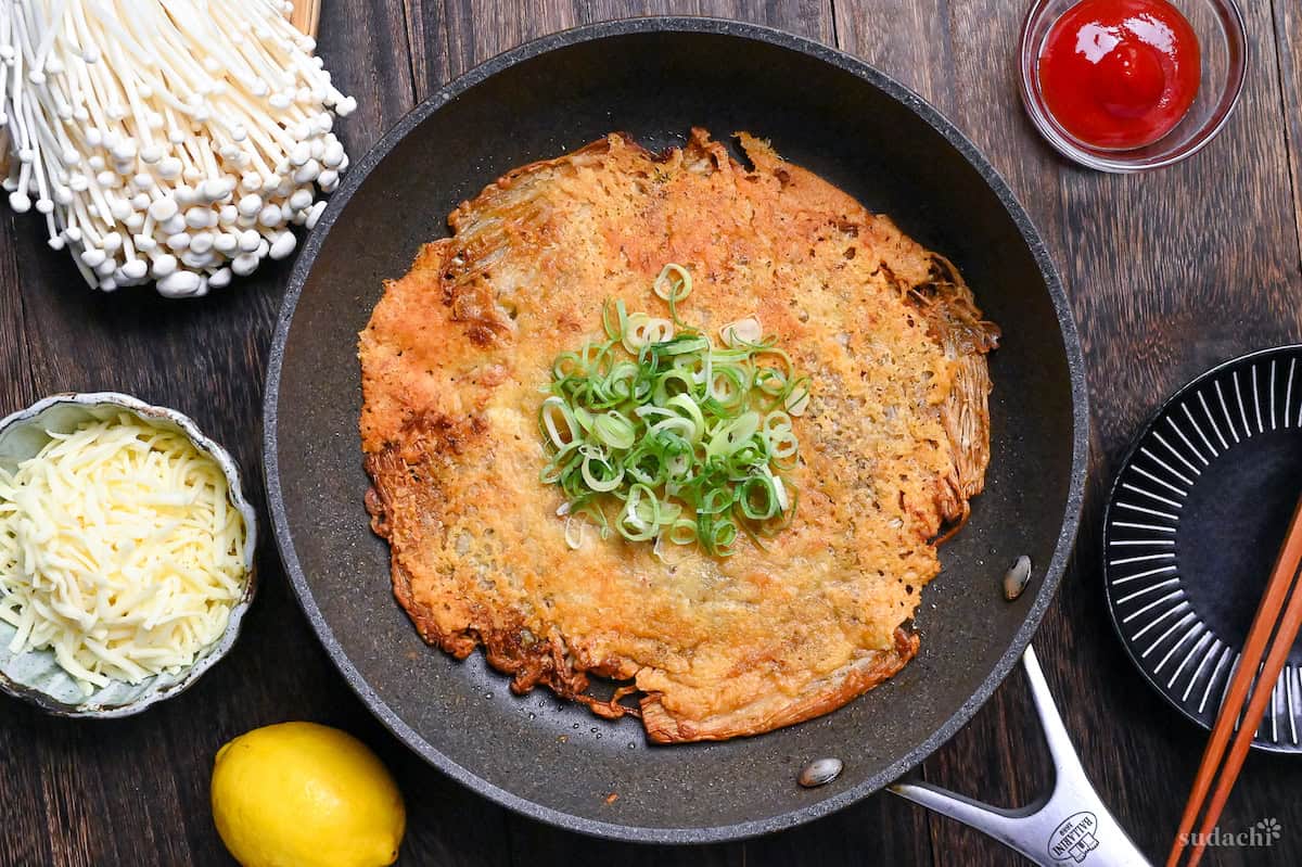 Crispy enoki cheese in a frying pan topped with chopped green onions next to a small bowl of ketchup, shredded cheese, a black plate, wooden chopsticks, enoki mushrooms and a lemon top down view