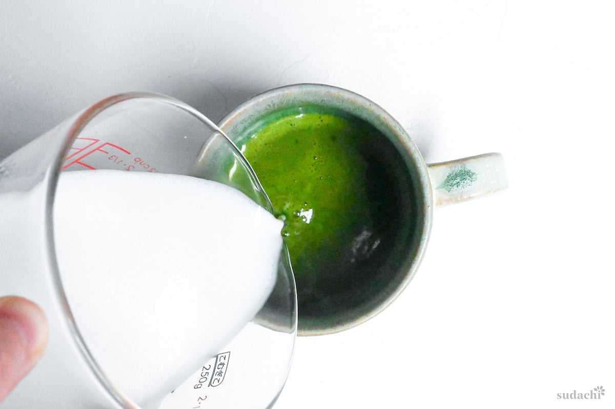 pouring frothed milk over whisked matcha into a teal ceramic cup