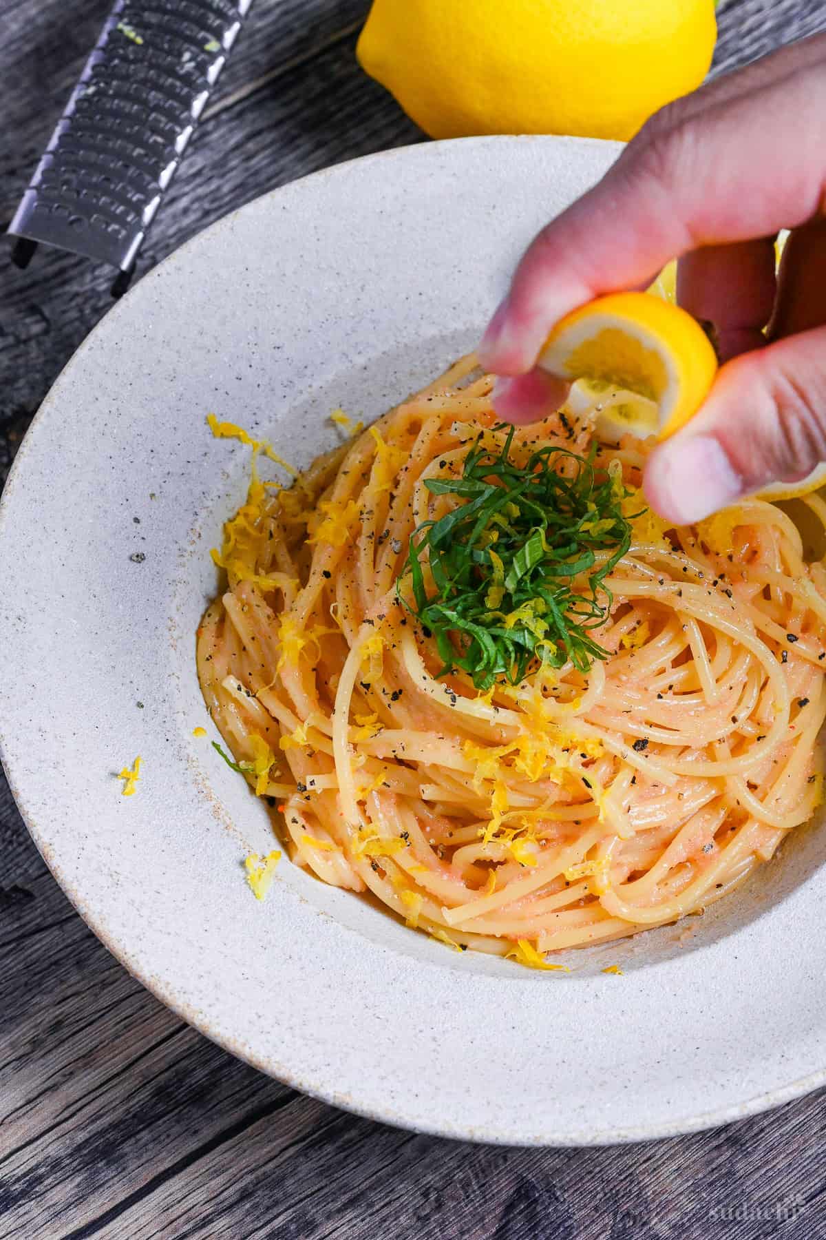 a hand squeezing a piece of lemon over mentaiko pasta in an off-white dish