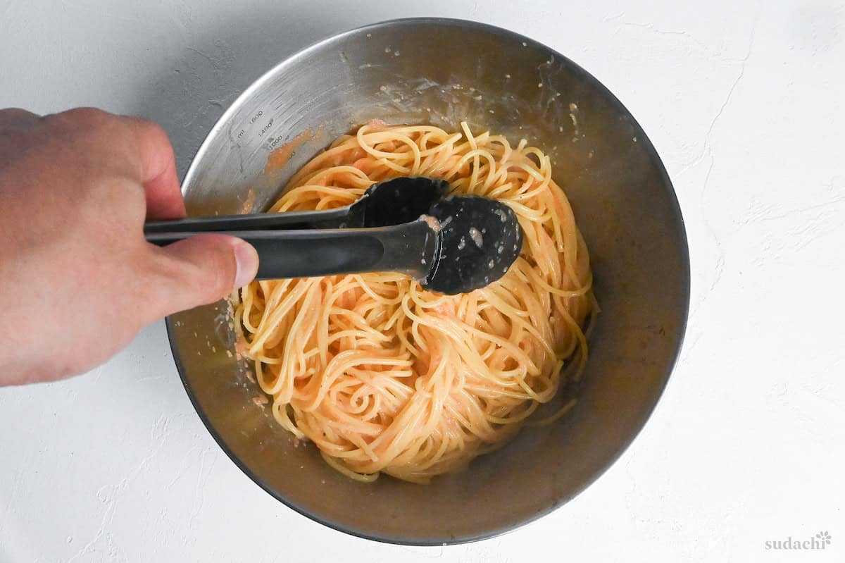Mentaiko pasta sauce and freshly cooked spaghetti in a steel mixing bowl being mixed with tongs to coat every strand