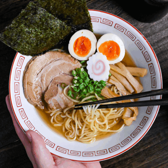 Homemade shoyu ramen in a white and red ramen bowl topped with pork chashu, ramen eggs, narutomaki, nori, chopped green onions and menma with noodles lifted with black chopsticks