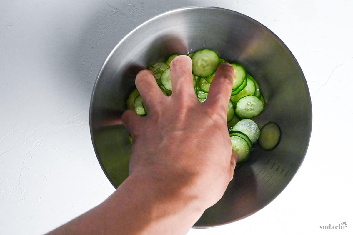 a hand massaging salted cucumber in a steel mixing bowl