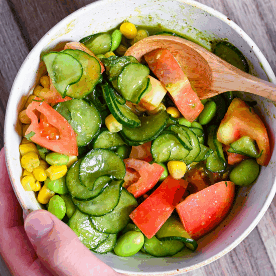 a hand holding a bowl filled with summer vegetable salad and shiso dressing with a wooden spoon close up