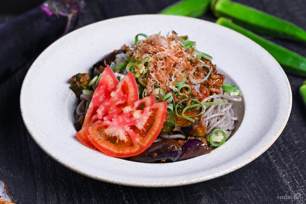 Soba noodle salad made with summer vegetables and miso meat sauce in an off-white bowl topped with chopped green onions, bonito flakes and sliced tomato on a black surface