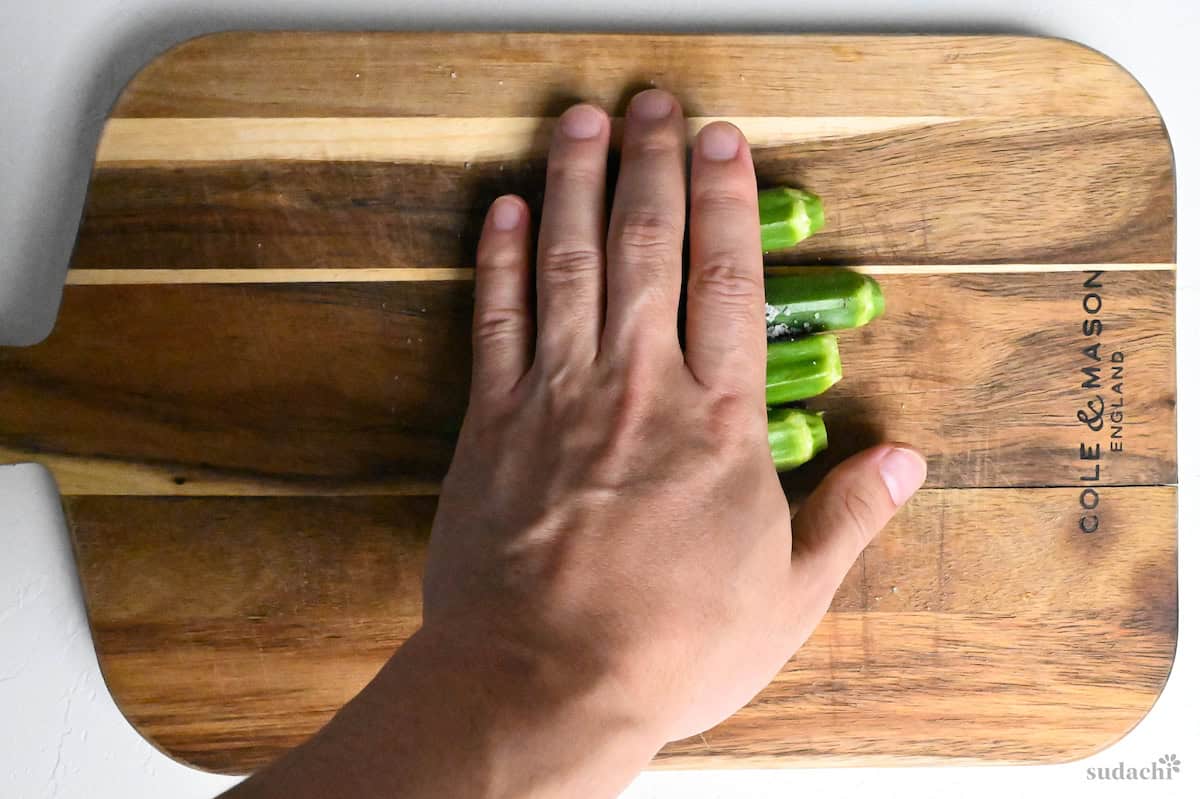 rolling 4 okra with salt on a wooden cutting board to remove fibers