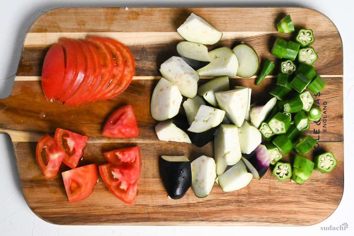 sliced tomato, roughly cut eggplant and okra on a wooden cutting board