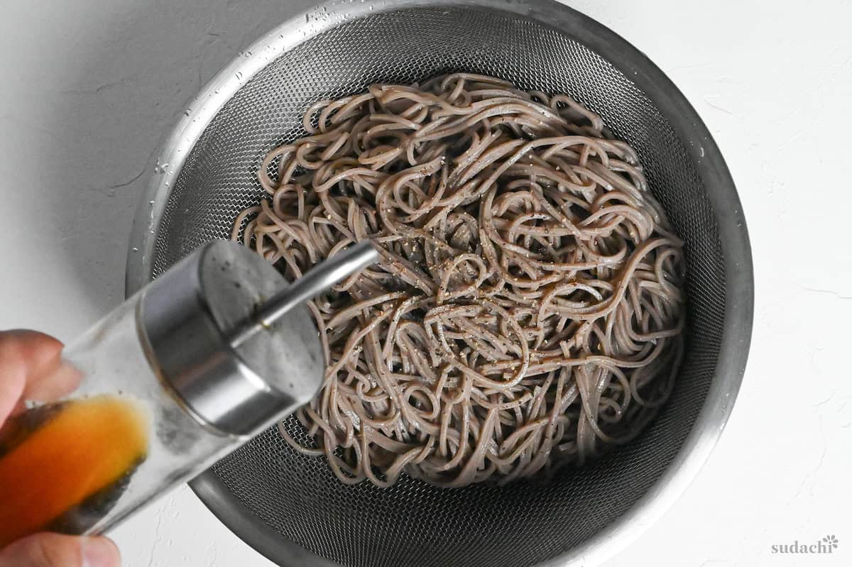 cooked soba noodles in a fine mesh sieve over a bowl sprinkled with ground black pepper and being drizzled with toasted sesame oil
