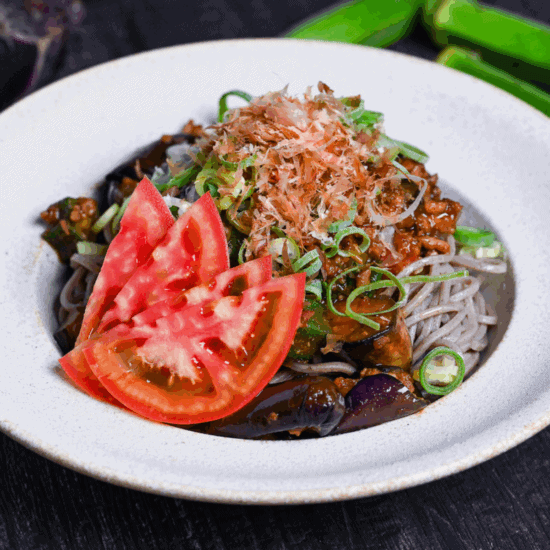 Soba noodle salad made with summer vegetables and miso meat sauce in an off-white bowl topped with chopped green onions, bonito flakes and sliced tomato on a black surface