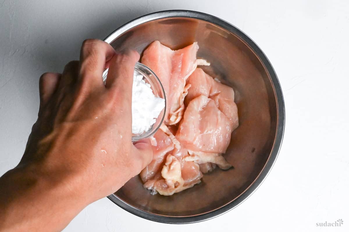 pouring starch over chicken breast pieces in a metal mixing bowl