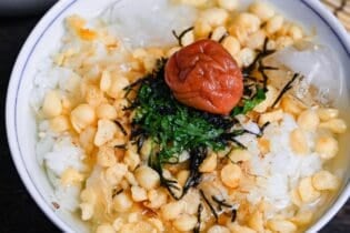 Chilled ume chazuke topped with tempura flakes, bonito flakes, kizami nori, shredded shiso leaves and one umeboshi in a white and blue flower-design bowl