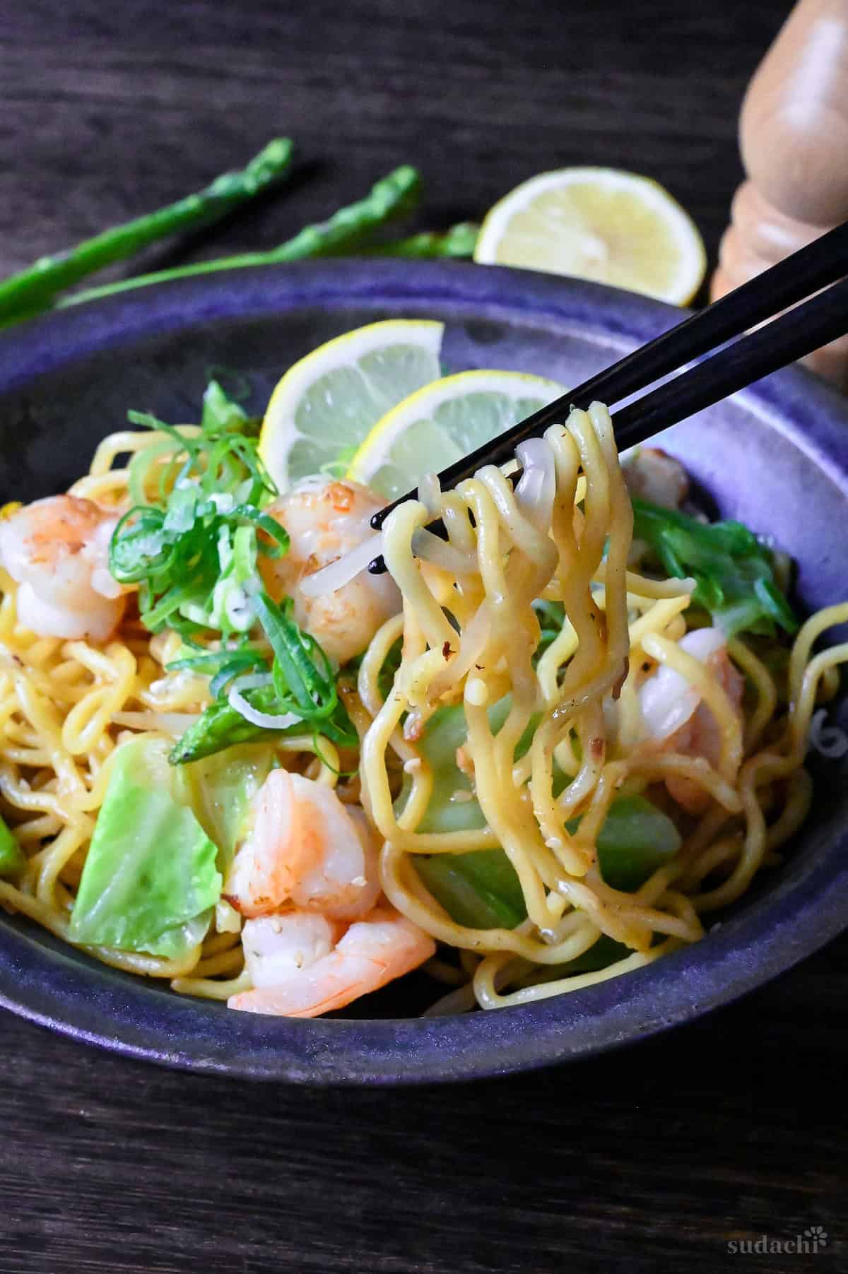 Shio Lemon Yakisoba in a blue/black iridescent bowl with black chopsticks holding up noodles