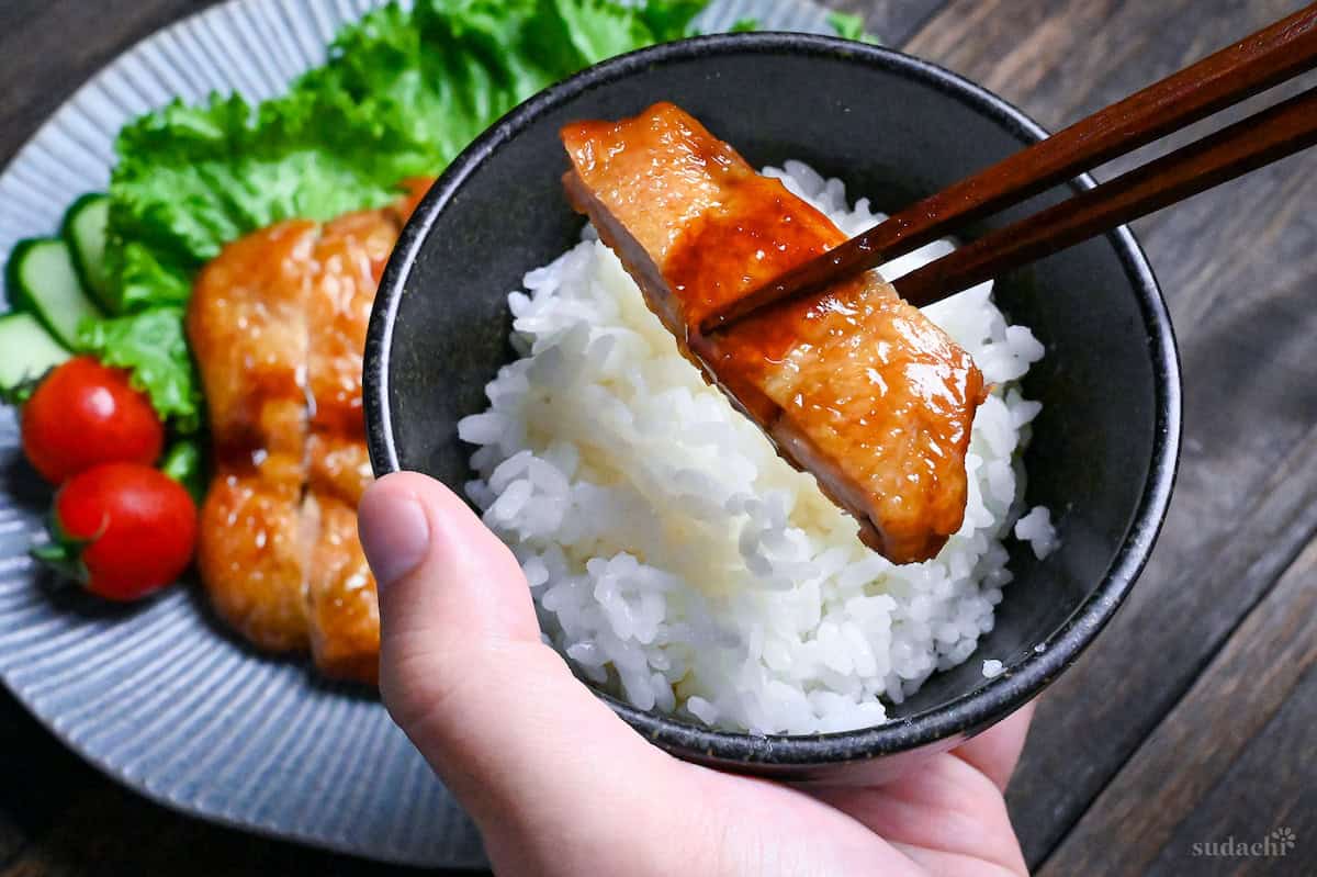 Wooden chopsticks holding a piece of teriyaki chicken over rice in a small black Japanese rice bowl with a blue plate in the background