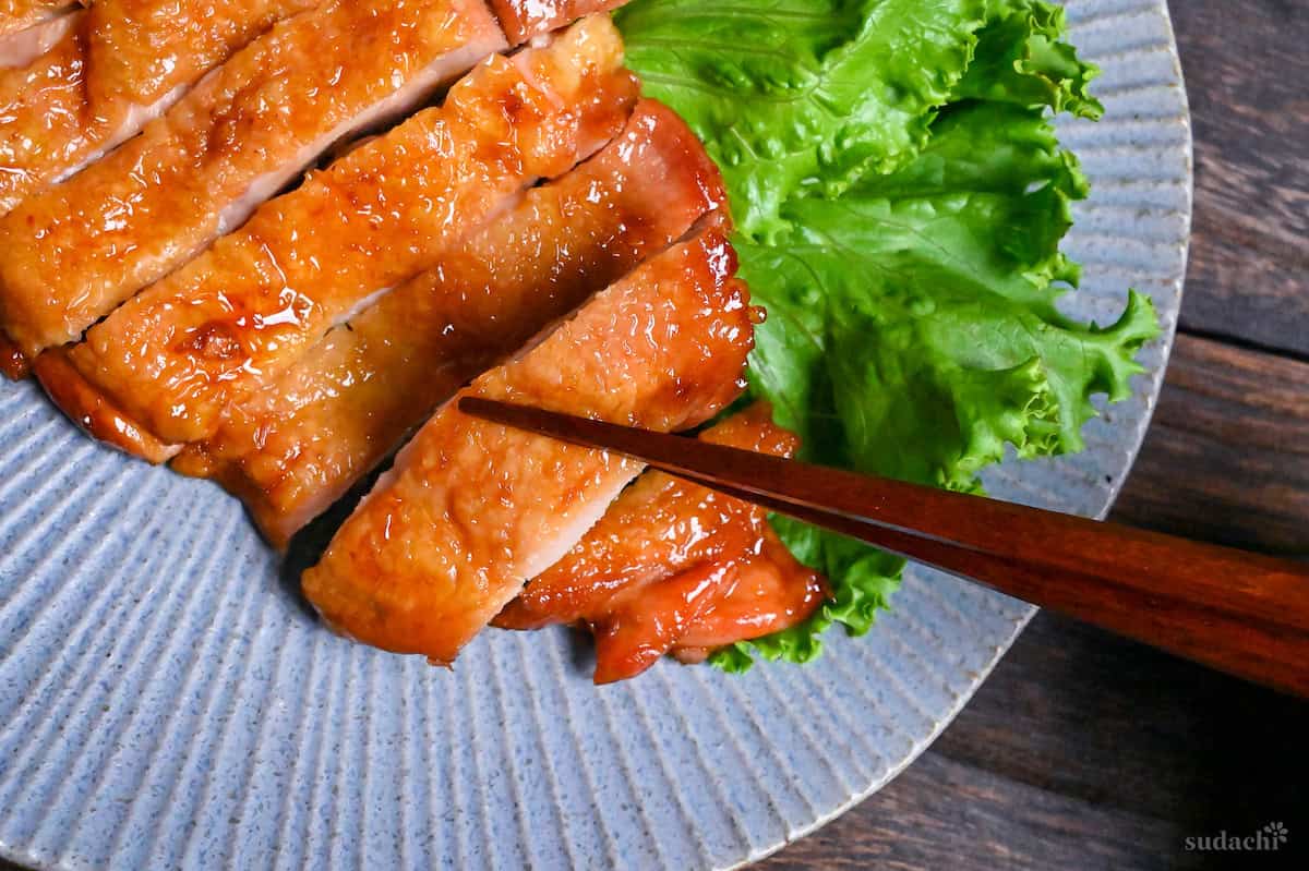 A close up of wooden chopsticks picking up a piece of teriyaki chicken from a blue plate