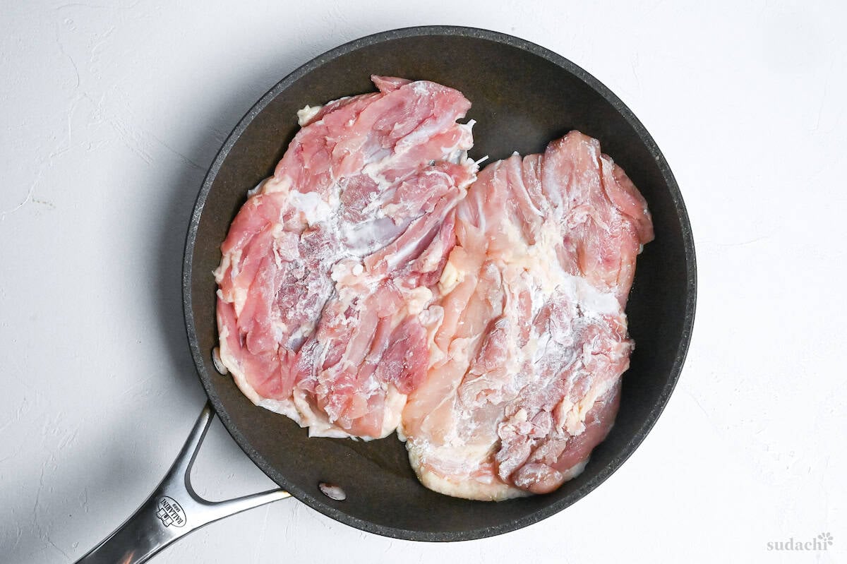Two skin-on boneless chicken thighs in a cold frying pan on a white background
