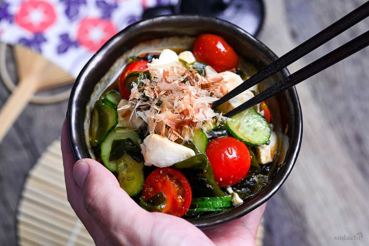 Japanese wakame salad made with wakame seaweed, mini tomatoes, cucumber and silken tofu served in a mottled gray bowl with dark brown edge held in a hand with black chopsticks picking up from the middle