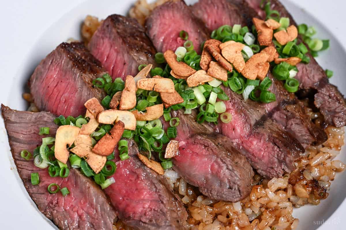 close up of garlic fried rice topped with rare pan-fried beef steak slices, chopped green onions and crispy garlic chips in a white dish