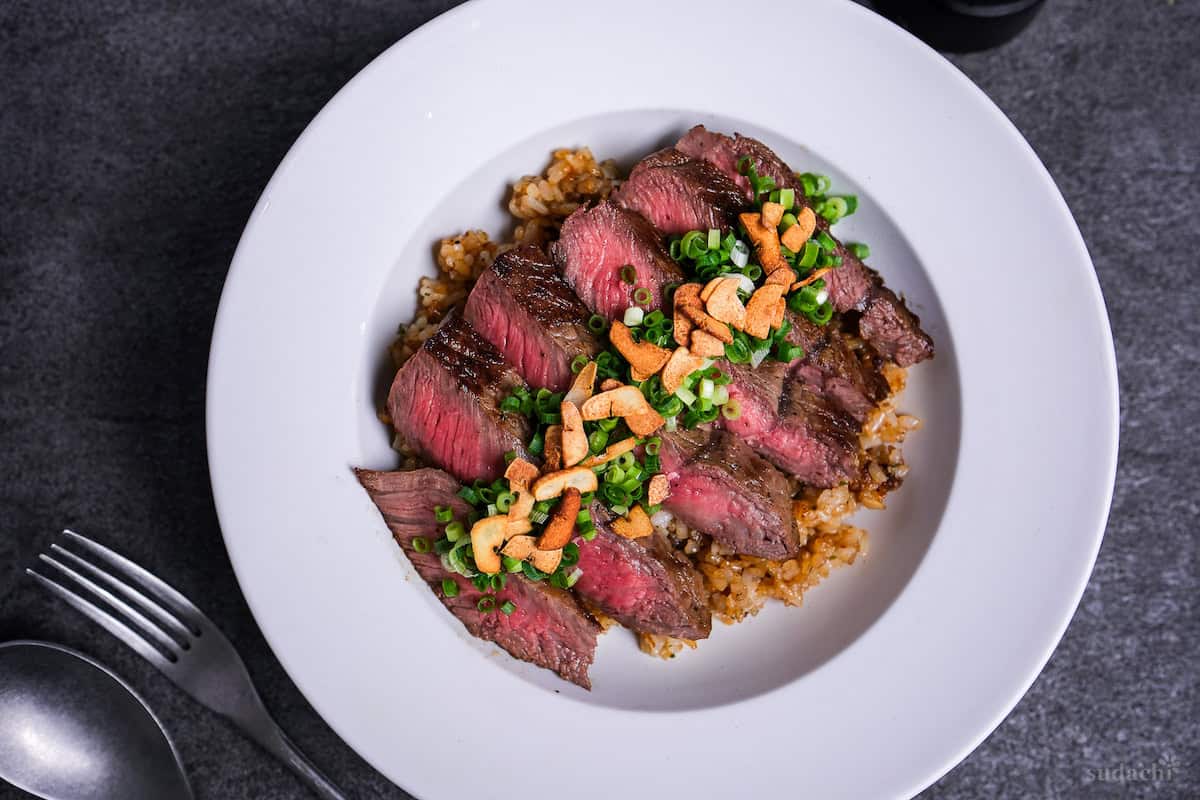 garlic fried rice topped with rare pan-fried beef steak slices, chopped green onions and crispy garlic chips in a white dish top down