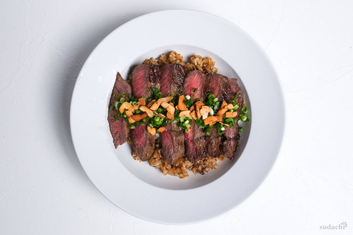garlic fried rice topped with rare pan-fried beef steak slices, chopped green onions and crispy garlic chips in a white dish
