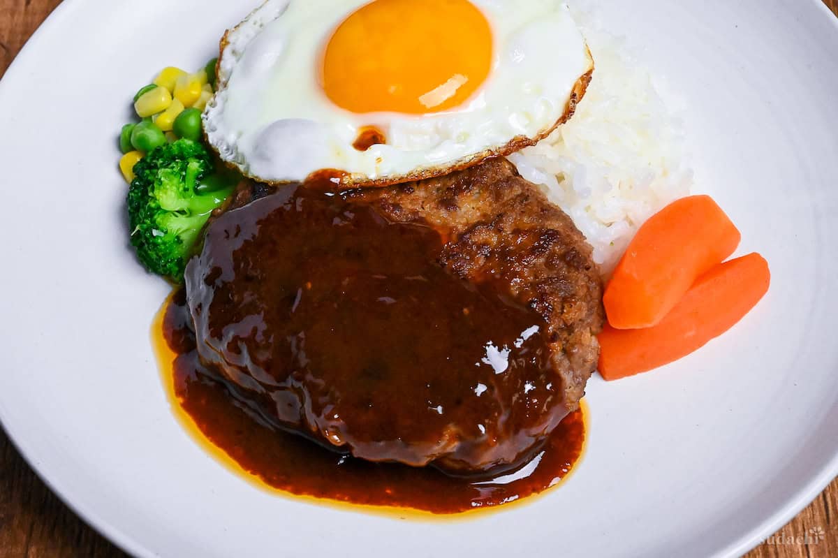 Japanese hamburg steak (hambagu) coated with homemade sauce on a white plate served with rice, vegetables and a fried egg